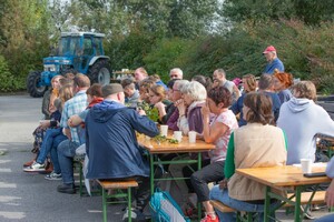 Eine Gruppe von Menschen bei einem Ouvertura-Hoffest auf Heurigentischen in der Sonne sitzend.